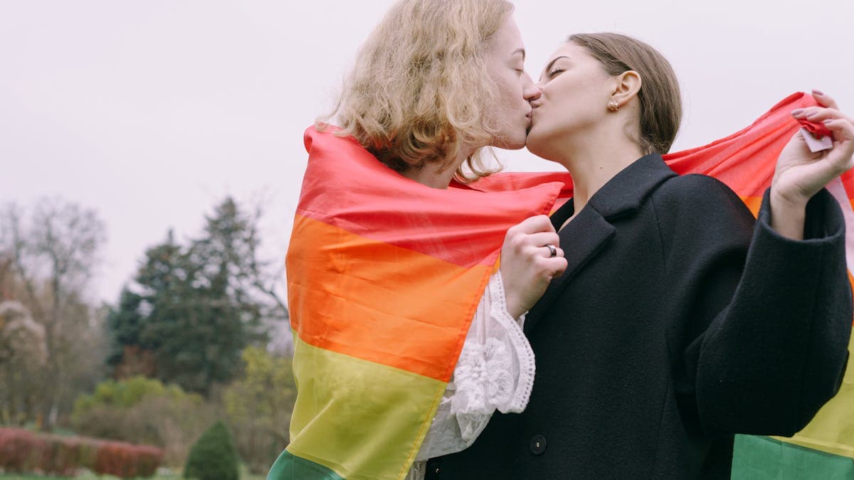 LGBTQ+ couple holding a rainbow pride flag and kissing outdoors, celebrating queer love
