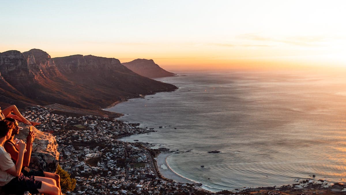 Young tourists watching the sunset while sitting on a mountain above Cape Town, with the coastline glowing below